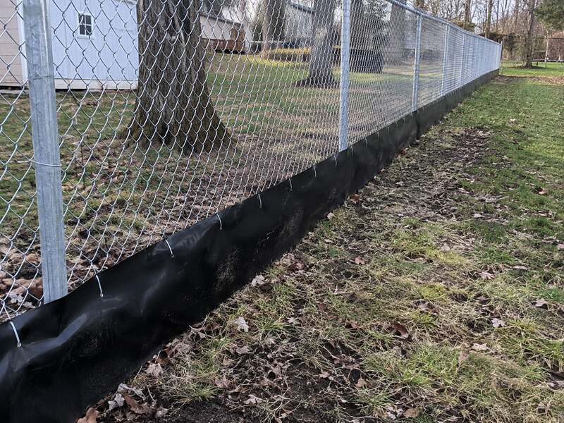 Black HDPE under fence barrier along a chain link fence in an open yard with a shed
