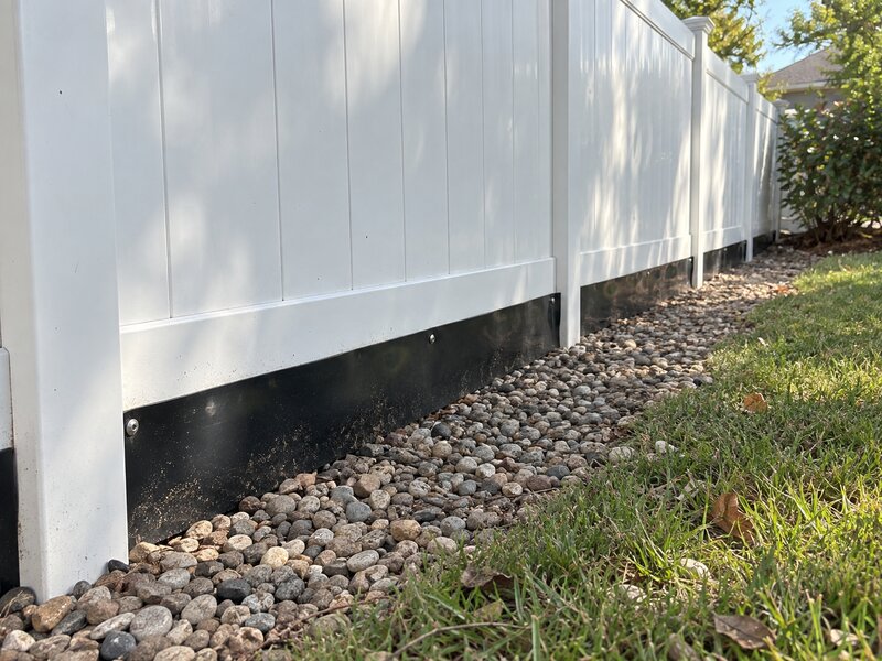 Black HDPE dig barrier installed at the base of a white vinyl fence over a river rock bed
