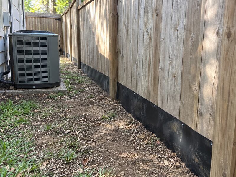 Black HDPE dig barrier along a wood privacy fence in a narrow side yard near an AC unit
