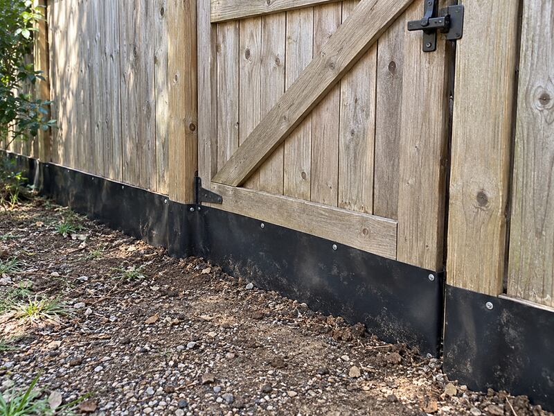 Close-up of HDPE dig barrier installed along a wood fence gate and gravel path
